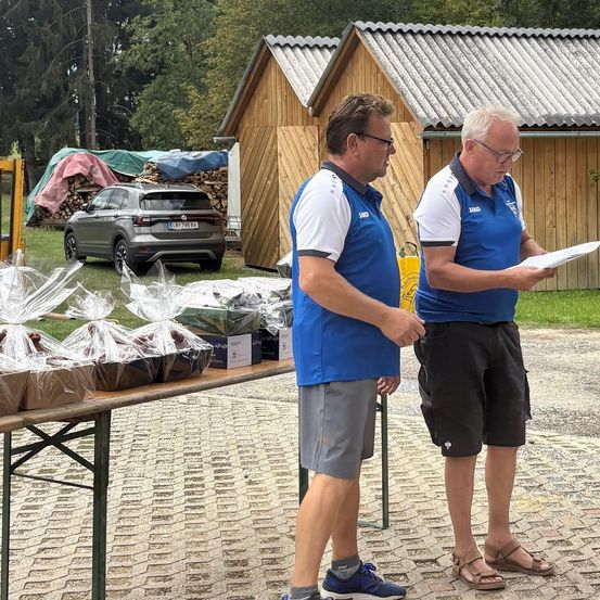 Zwei Männer in blauen Hemden und Shorts stehen neben einem Tisch mit verpackten Gegenständen, möglicherweise bei einer Outdoor-Veranstaltung. Hinter ihnen befinden sich Holzstrukturen und ein Auto.