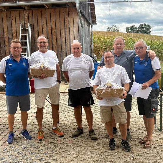 Fünf Männer stehen vor einer Scheune und halten Geschenkkörbe in den Händen. Sie sind lässig gekleidet und scheinen für ein Foto zu posieren. Dahinter befindet sich ein Maisfeld und eine Leiter, die an der Scheune gelehnt ist.