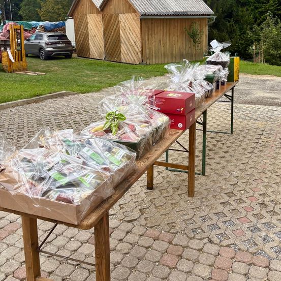 Ein langer Holztisch im Freien, bedeckt mit verschiedenen Lebensmitteln in Plastiktüten und Kisten, vor einem kleinen Holzgebäude aufgestellt.