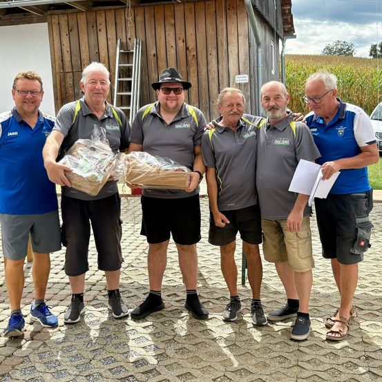 Fünf Männer stehen vor einem Holzgebäude, zwei von ihnen halten verpackte Geschenke. Sie tragen alle graue Shirts mit Logos, und einer der Männer trägt einen Hut.
