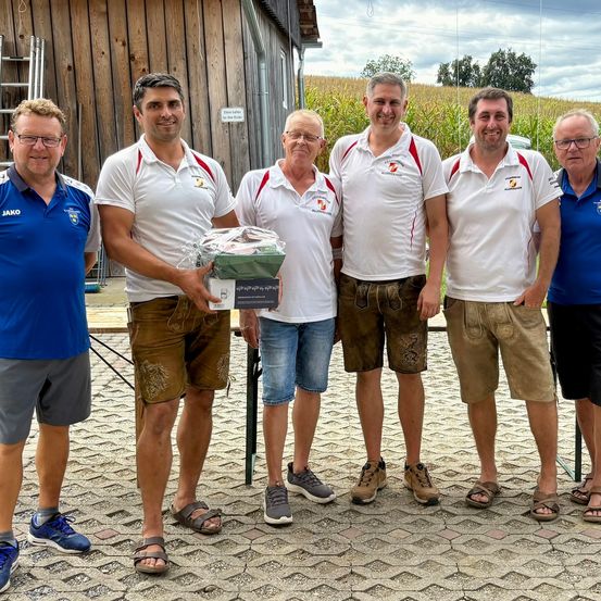 Fünf Männer stehen lächelnd in einer Reihe vor einer Holzscheune mit einem Maisfeld im Hintergrund. Sie tragen passende weiße Poloshirts mit roten und weißen Akzenten. Der Mann in der Mitte hält eine Schachtel.
