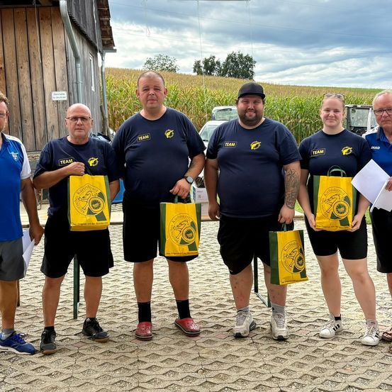 Eine Gruppe von sechs Personen, alle in blauen T-Shirts mit einem Logo, steht vor einer Scheune und einem Maisfeld und hält gelbe Taschen.
