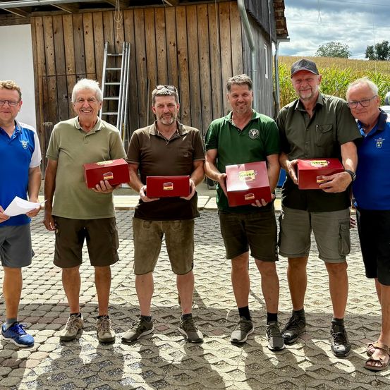 Fünf Männer stehen in einer Reihe, halten rote Boxen und posieren lächelnd für ein Foto vor einem Holzgebäude.