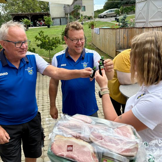 Zwei Männer in blauen Shirts und eine Frau in einem weißen Shirt stehen um einen Tisch mit Fleisch in Plastiktüten. Sie halten kleine Flaschen und lächeln.