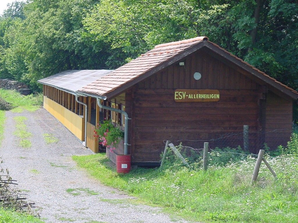 Ein hölzernes Gebäude mit einem roten Dach und dem Schild ESV-Alleheiligen steht auf einem Kiesweg. An der Seite befinden sich Pflanzen und Blumen.