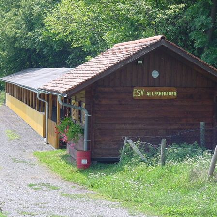 Ein hölzernes Gebäude mit einem roten Dach und dem Schild ESV-Alleheiligen steht auf einem Kiesweg. An der Seite befinden sich Pflanzen und Blumen.