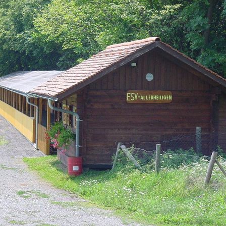 Ein Holzgebäude mit braunem Dach und dem Namen ESV-Allerheiligen steht an einer Kiesstraße.