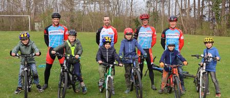A group of cyclists, including adults and children, pose for a photo on a grassy field with leafless trees in the background.