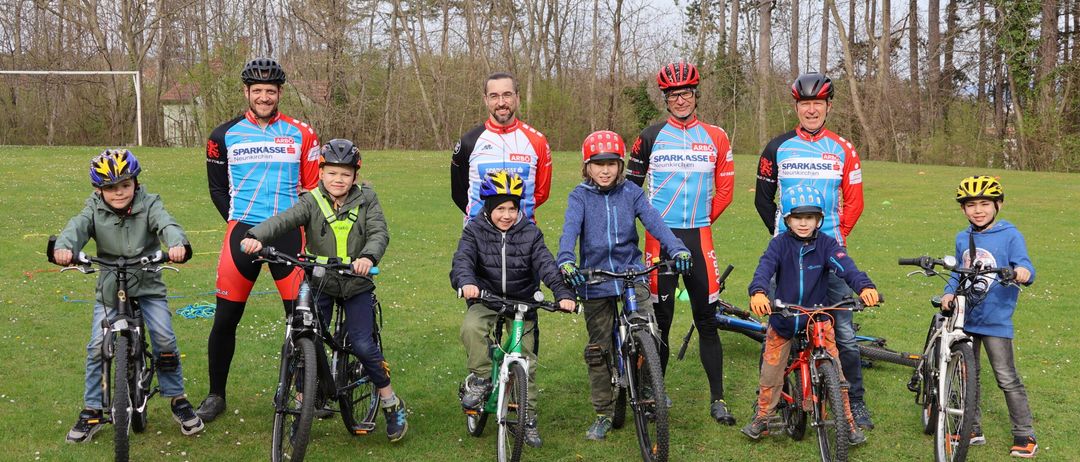 Eine Gruppe von Radfahrern, einschließlich Erwachsener und Kinder, posiert für ein Foto auf einer grasbewachsenen Wiese mit laublosen Bäumen im Hintergrund.