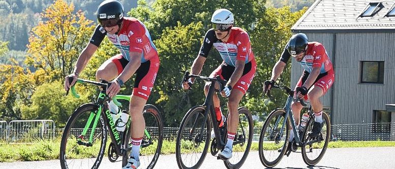 Drei Radfahrer in roten Radsporttrikots fahren auf einer Straße mit Bergen und einem Gebäude mit Solarmodulen im Hintergrund.