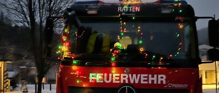 Ein Feuerwehrauto mit der Aufschrift 'Feuerwehr' vorne, geschmückt mit Weihnachtslichtern, auf einer verschneiten Straße geparkt.