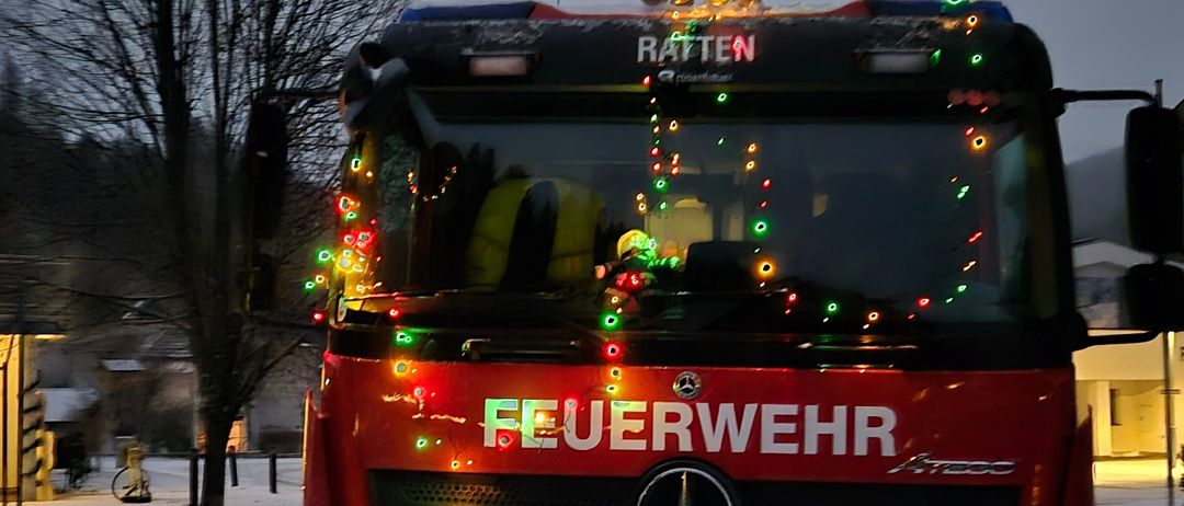 Ein Feuerwehrauto mit der Aufschrift 'Feuerwehr' vorne, geschmückt mit Weihnachtslichtern, auf einer verschneiten Straße geparkt.