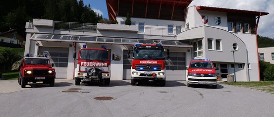 Drei Feuerwehrfahrzeuge stehen vor einer Feuerwehrstation mit rotem Dach und zwei Emblemen. Die Fahrzeuge haben blaue Lichter auf dem Dach.