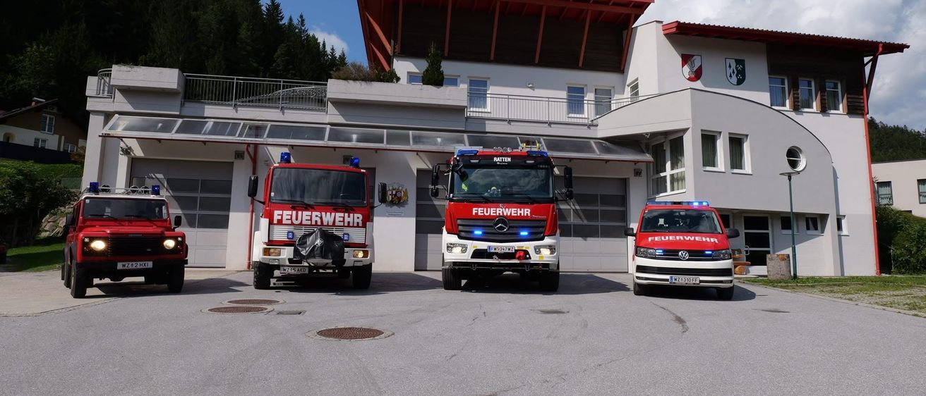 Drei Feuerwehrfahrzeuge stehen vor einer Feuerwehrstation mit rotem Dach und zwei Emblemen. Die Fahrzeuge haben blaue Lichter auf dem Dach.