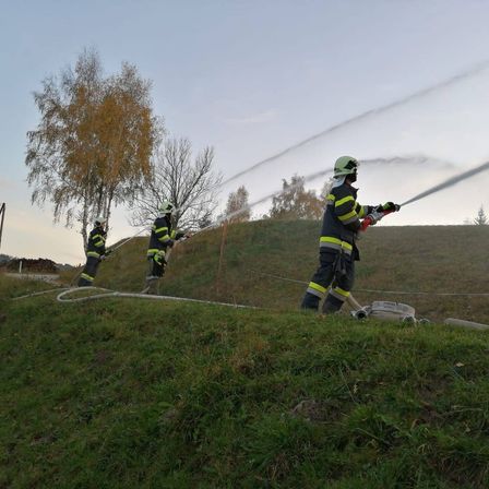 Drei Feuerwehrleute in gelben und schwarzen Uniformen sprühen Wasser aus einem Schlauch auf einen grasbewachsenen Hügel unter einem klaren Himmel.