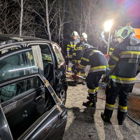 Feuerwehrleute in Schutzkleidung arbeiten nachts an einem beschädigten Auto und benutzen Werkzeuge und eine Taschenlampe.