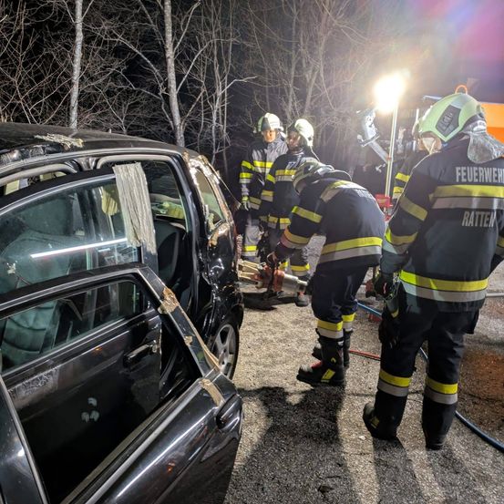 Feuerwehrleute in Schutzkleidung arbeiten nachts an einem beschädigten Auto und benutzen Werkzeuge und eine Taschenlampe.