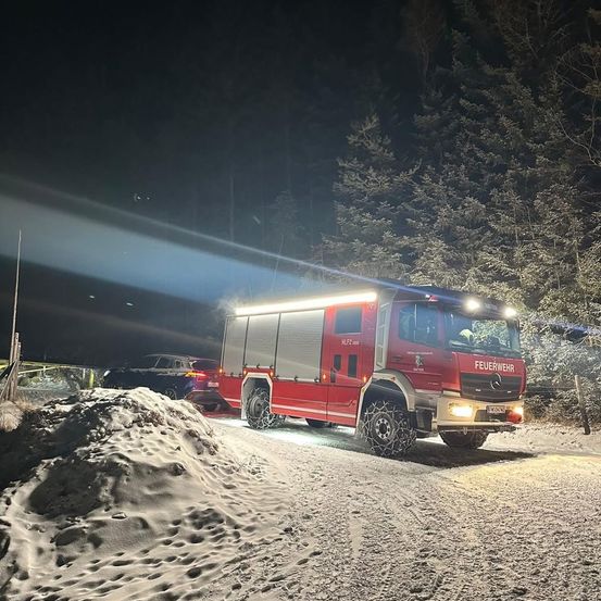 Ein roter Feuerwehrwagen mit eingeschalteten Scheinwerfern steht bei Nacht auf einer verschneiten Straße, mit einem Schneehaufen links und einem geparkten Auto dahinter.