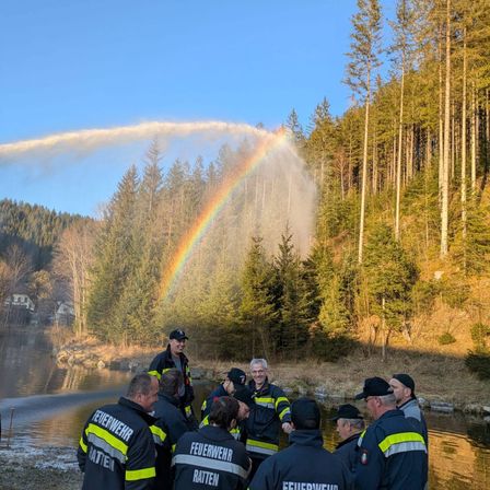 Eine Gruppe von Feuerwehrleuten in Uniform steht am Ufer eines Flusses, mit einem Regenbogen am Himmel über ihnen.