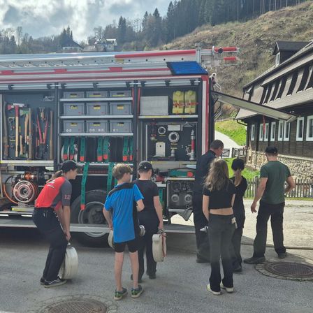Eine Gruppe von Menschen, einschließlich Kindern, steht um einen Feuerwehrwagen mit offenen Türen herum, der verschiedene Feuerwehrausrüstung zeigt. Die Szene spielt sich in einer ländlichen Gegend mit einem Haus und einem Hügel im Hintergrund ab.
