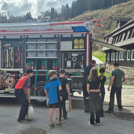 Eine Gruppe von Menschen, einschließlich Kindern, steht um einen Feuerwehrwagen mit offenen Türen herum, der verschiedene Feuerwehrausrüstung zeigt. Die Szene spielt sich in einer ländlichen Gegend mit einem Haus und einem Hügel im Hintergrund ab.