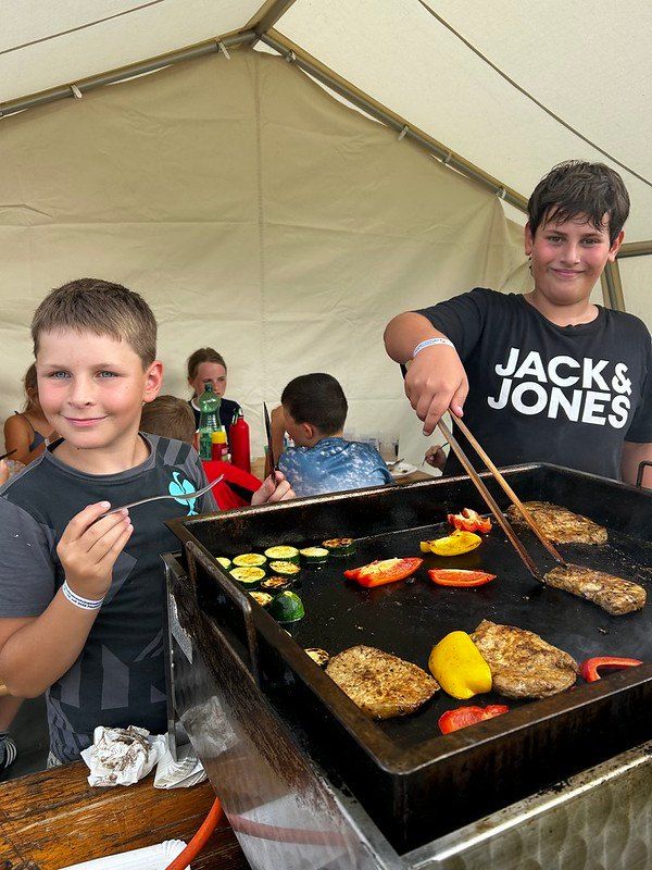 Zwei Jungen grillen Gemüse und Fleisch auf einem Grill unter einem Zelt. Ein Junge hält eine Gabel und der andere benutzt eine Zange.
