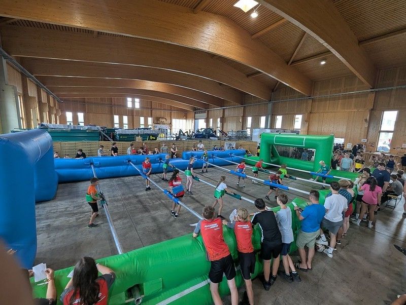 Eine Gruppe von Menschen spielt ein Spiel in einer großen Indoor-Anlage mit aufblasbaren Hindernissen. Einige nehmen aktiv teil, während andere von den Seitenlinien aus zuschauen. Die Anlage hat Holzwände und eine hohe Decke.