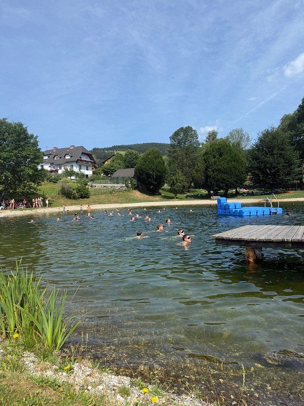Eine Gruppe von Menschen schwimmt in einem See mit einem hölzernen Steg und blauen Sprungbrettern im Hintergrund.