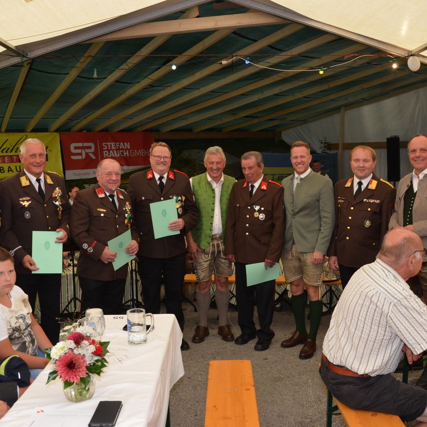 Eine Gruppe von Männern in Militäruniformen und traditioneller bayerischer Kleidung steht in einem Zelt, hält Urkunden und lächelt für ein Foto. Ein Tisch mit Blumen und ein Junge, der in der Nähe sitzt.