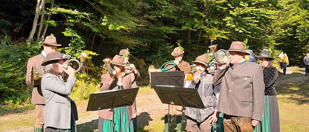 Eine Gruppe von Menschen in traditionellen Kostümen steht in einem Wald und spielt Blechblasinstrumente.