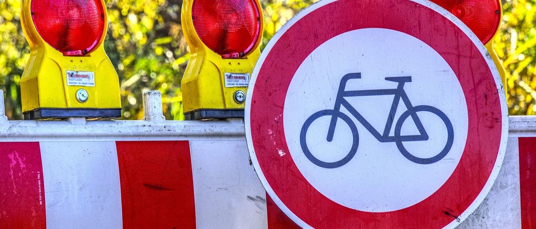 Ein Fahrradschild auf einer Absperrung mit roter Warnleuchte, im Freien in der Nähe von Bäumen mit gelben Blättern.