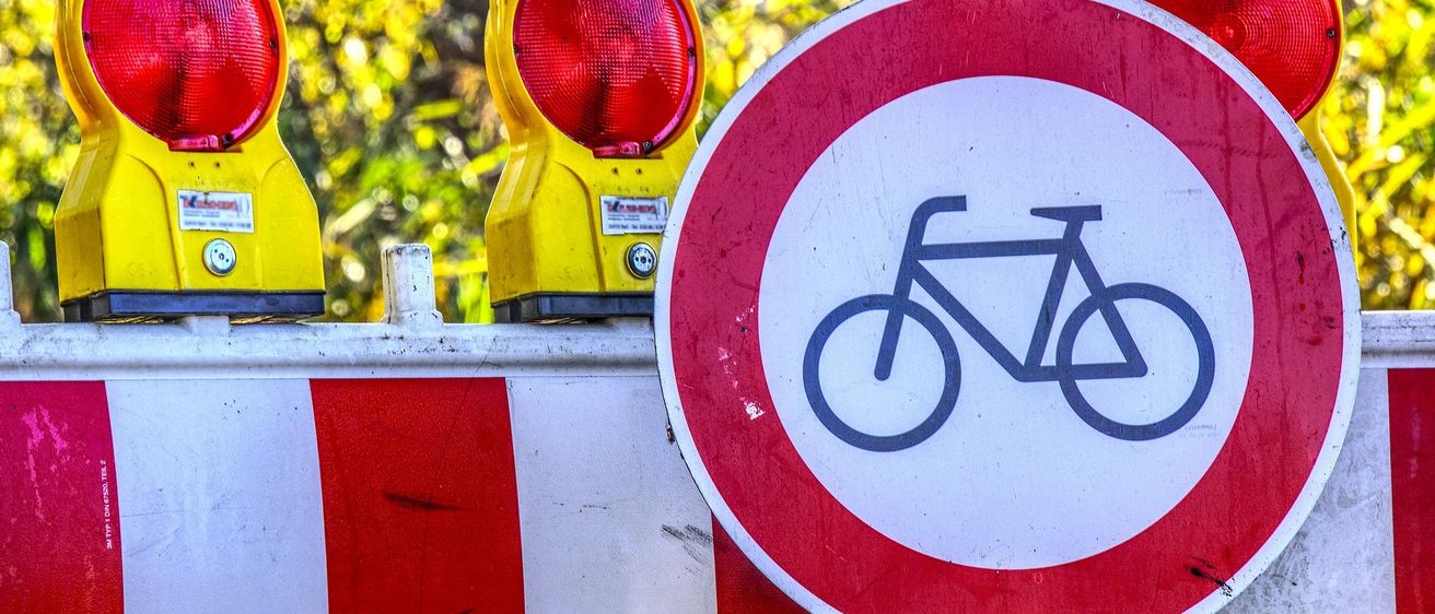 Ein Fahrradschild auf einer Absperrung mit roter Warnleuchte, im Freien in der Nähe von Bäumen mit gelben Blättern.