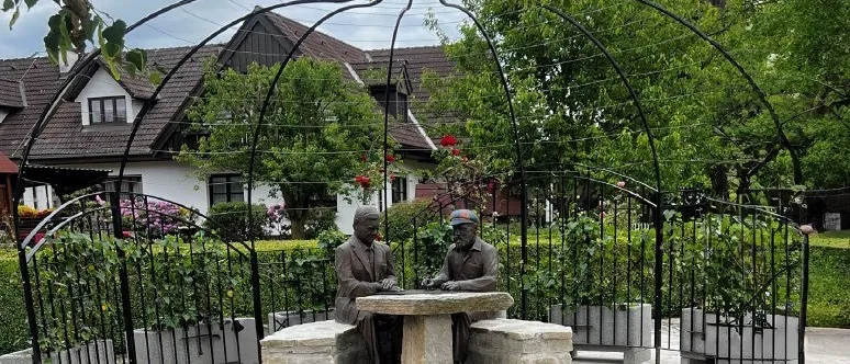 Zwei Statuen von Männern sitzen an einem steinernen Tisch in einem Garten mit einem Haus und einem Zaun im Hintergrund.