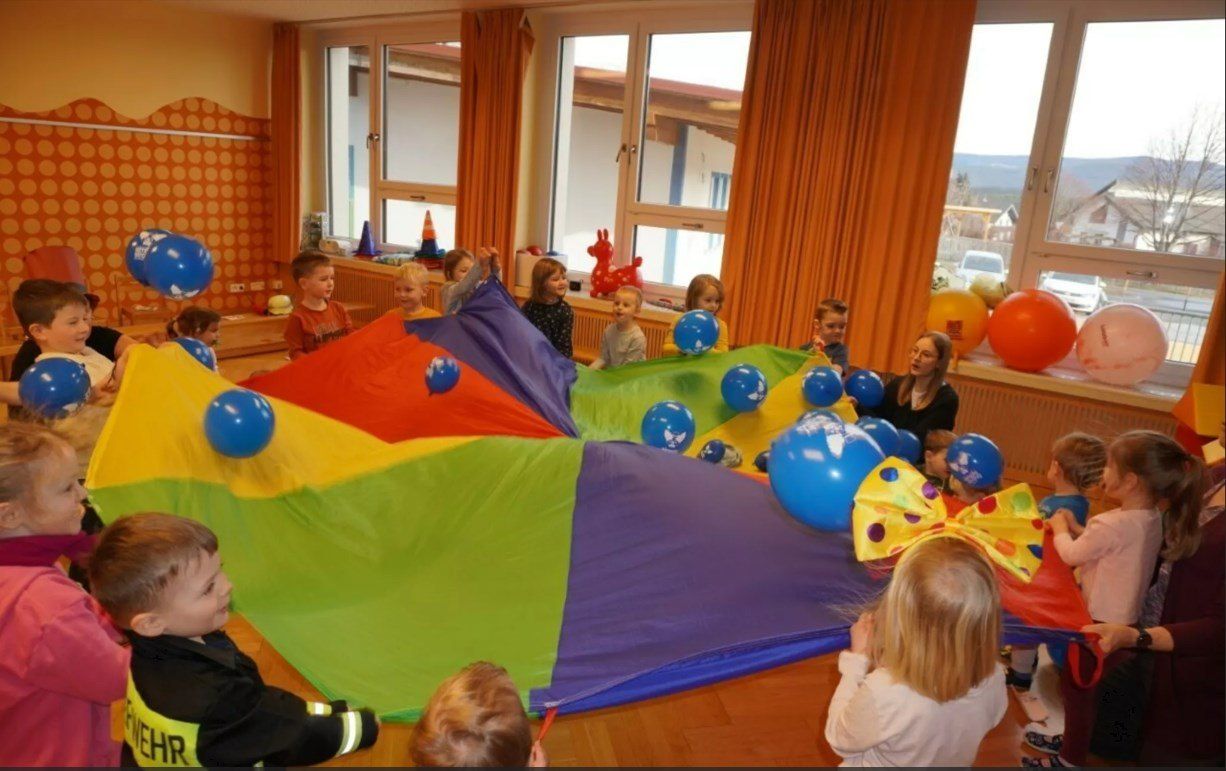 Mehrere Kinder spielen mit bunten Luftballons in einem Raum mit großen Fenstern. Ein Erwachsener beaufsichtigt sie.