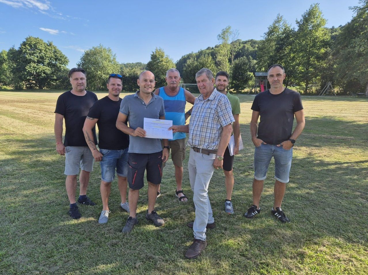 Eine Gruppe von Männern steht auf einer Wiese. Ein Mann hält ein Blatt Papier, möglicherweise ein Zertifikat, während die anderen zusehen. Sie sind lässig gekleidet und im Hintergrund befinden sich ein Spielplatz und Bäume.