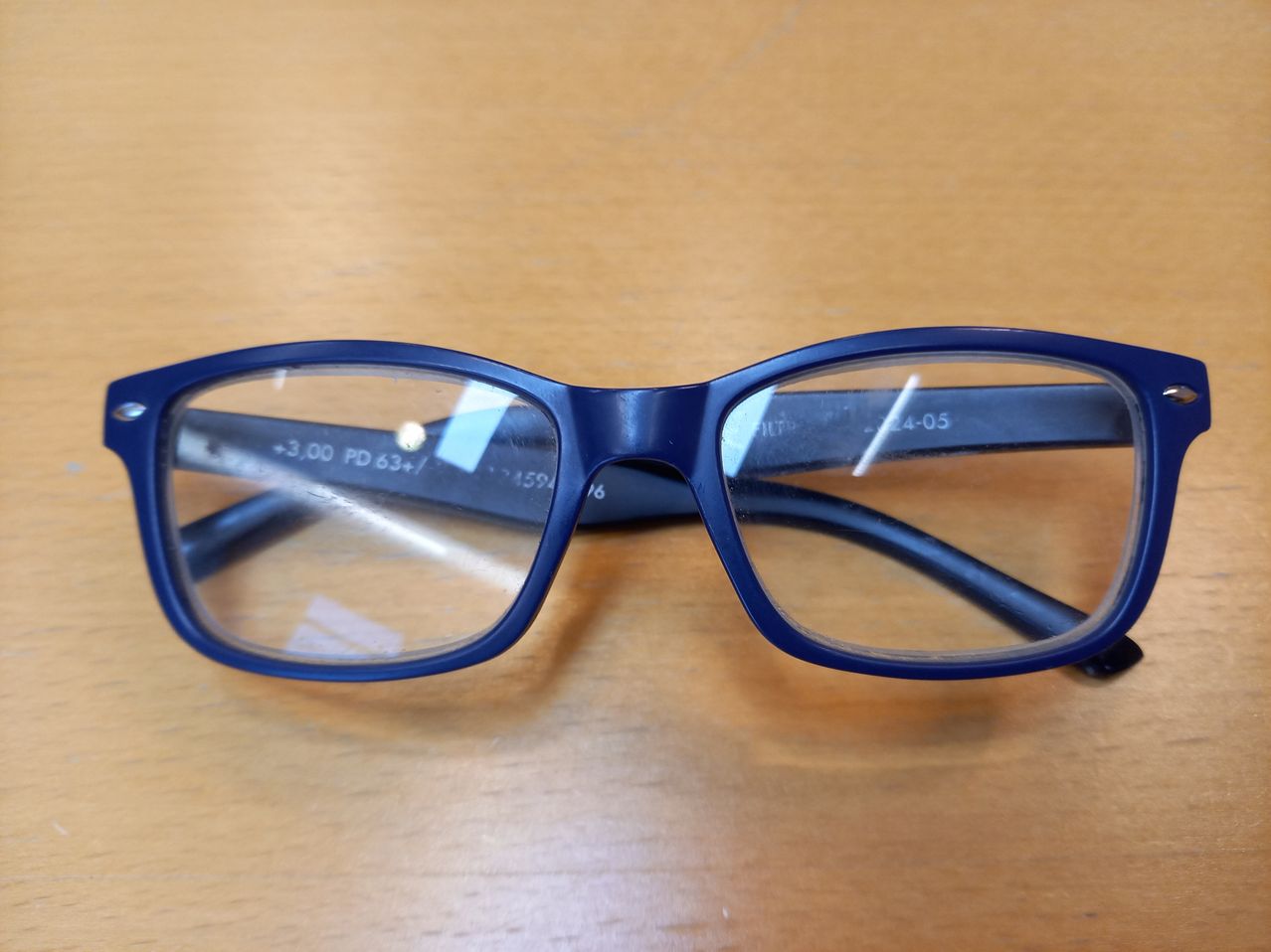 Ein Paar blauer Brille mit klaren Gläsern, beschriftet mit einer Sehstärke von +3.00 PD 63+. Die Gläser reflektieren eine hölzerne Oberfläche.