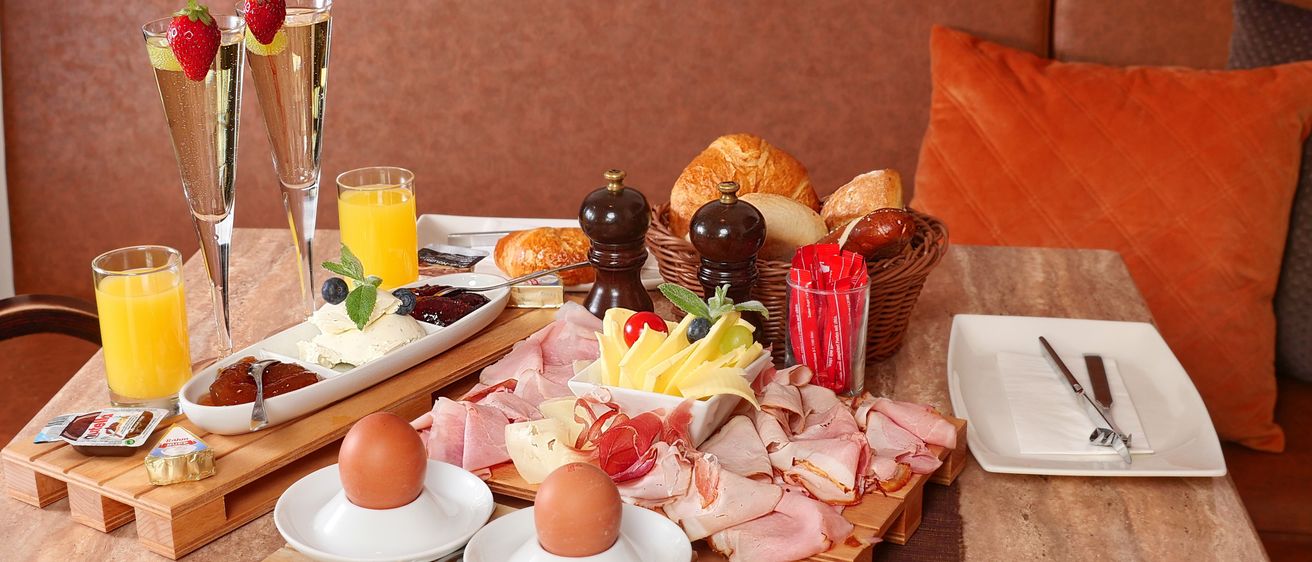 Ein Frühstückstisch mit Eiern, Fleisch, Käse, Obst, Brot und Getränken. Ein Korb mit Brot und Gewürzen in der Nähe.