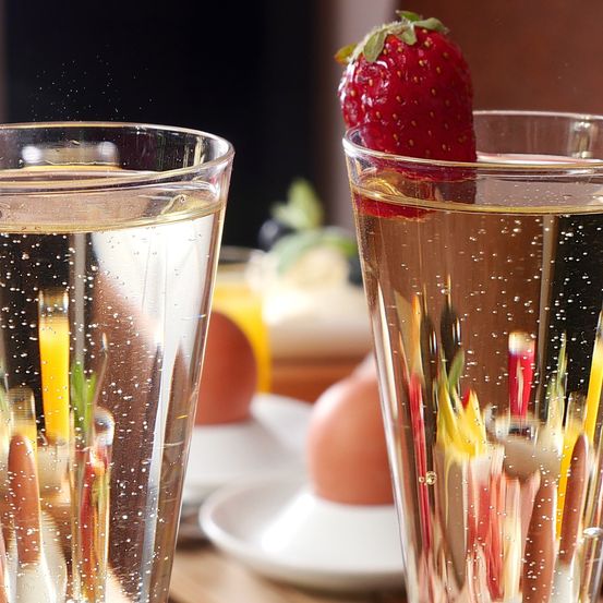Zwei Gläser mit prickelndem Champagner, obenauf eine Erdbeere, und ein Teller mit Eiern im Hintergrund.