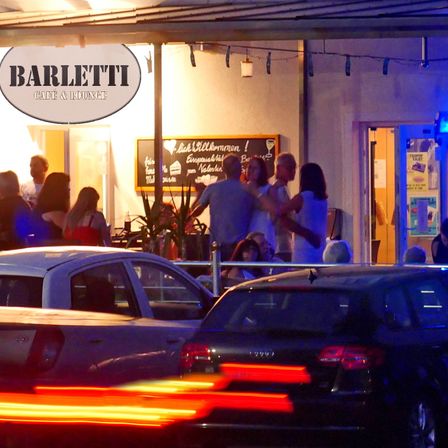 Im Barletti Cafe & Lounge stehen eine Gruppe von Menschen draußen. Vor dem Gebäude sind Autos geparkt und der Bereich ist von blauen Lichtern beleuchtet.
