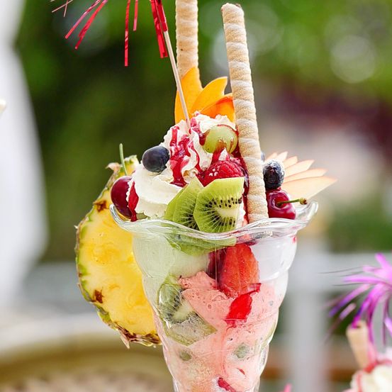 Ein Obstsundae in einem Glasbecher, gekrönt mit Schlagsahne, roter Soße und verschiedenen Früchten, mit einem Strohhalm und einer Ananasscheibe.