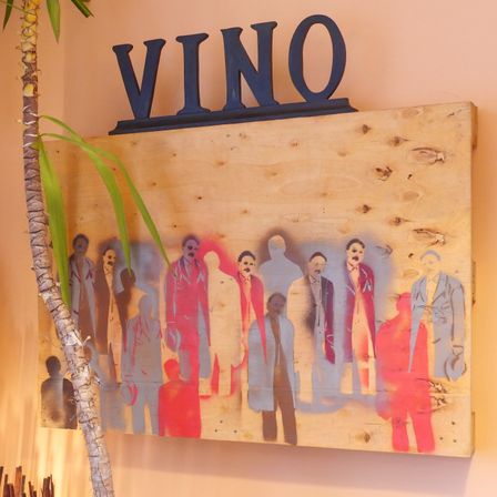 Ein Holzschild mit dem Wort 'VINO' darauf zeigt ein Gemälde von Männern in Anzügen, die in einer Reihe stehen.