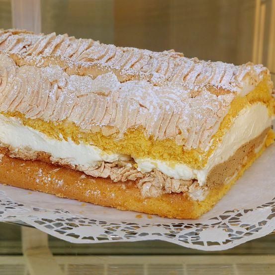 Ein gerollter Kuchen mit Schichten aus gelbem Biskuit, Sahne und Baiser. Der Kuchen ist mit Puderzucker bestreut und auf einem Spitzendeckchen serviert.