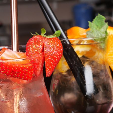 Zwei Cocktails in Gläsern mit Strohhalmen, einer mit einer Erdbeere und Eis, der andere mit Orangenscheiben und Minze. Ein schwarzer Strohhalm erstreckt sich vom Orangencocktail.