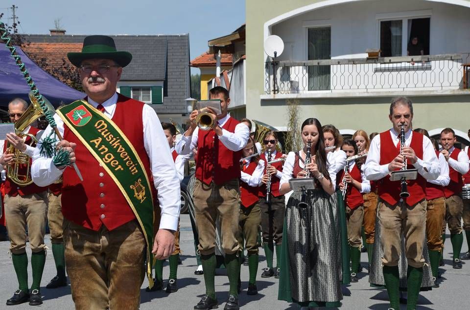 Eine Gruppe von Musikern in traditioneller Kleidung, einschließlich eines Mannes mit einem grünen Band, marschiert in einem Umzug. Hinter ihnen ist ein Gebäude mit Balkon und Satellitenschüssel zu sehen.