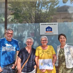 Vier Personen stehen vor einer Glaswand mit einem Schild, das 'SOZIAALVEREIN B37' besagt. Die Frau in der Mitte hält ein Zertifikat. Dahinter steht ein Baum und ein Gebäude.