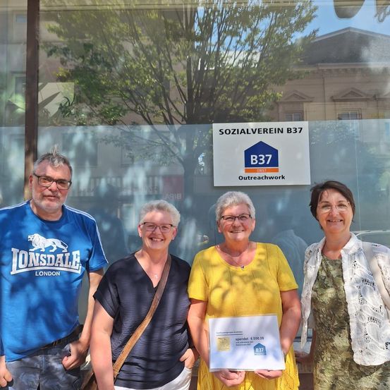 Vier Personen stehen vor einer Glaswand mit einem Schild, das 'SOZIAALVEREIN B37' besagt. Die Frau in der Mitte hält ein Zertifikat. Dahinter steht ein Baum und ein Gebäude.