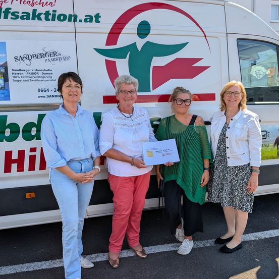 Vier Frauen stehen vor einem weißen Van mit rotem und grünem Logo. Sie lächeln und scheinen für ein Foto zu posieren. Die Frau in der Mitte hält ein Zertifikat.