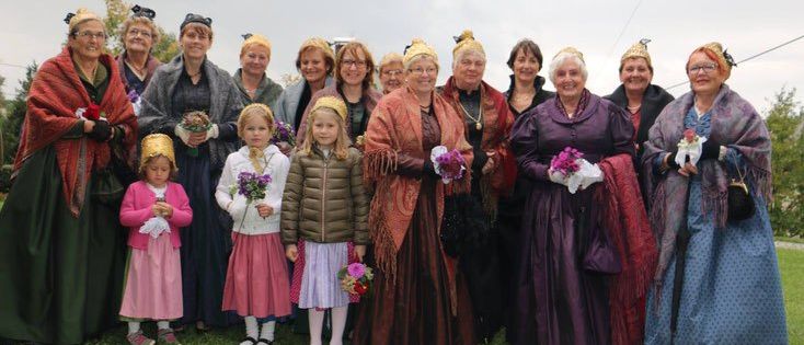 Eine Gruppe von Erwachsenen und Kindern in Kostümen posiert für ein Foto auf einer Rasenfläche. Die Erwachsenen tragen Vintage-Kleider und die Kinder halten Blumen. Sie lächeln und schauen in die Kamera. Der Hintergrund ist unscharf.