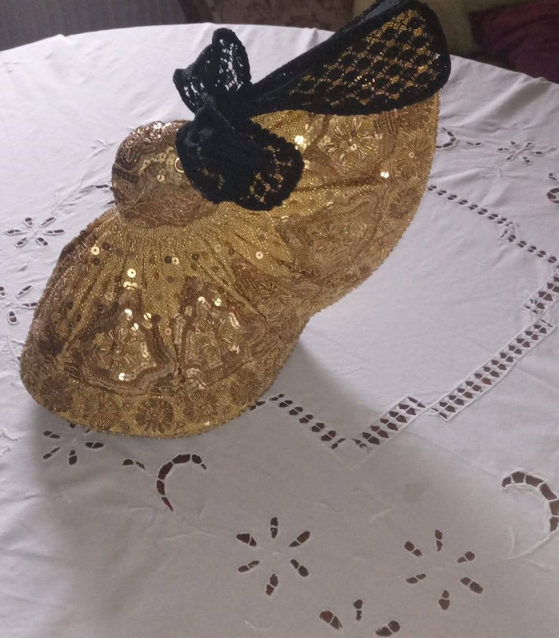 Ein goldener Absatzschuh mit einer schwarzen Spitzenschleife steht auf einem weißen Tuch. Der Schuh ist mit Pailletten verziert und hat ein Blumenmuster.
