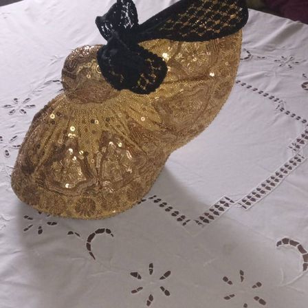 Ein goldener Absatzschuh mit einer schwarzen Spitzenschleife steht auf einem weißen Tuch. Der Schuh ist mit Pailletten verziert und hat ein Blumenmuster.
